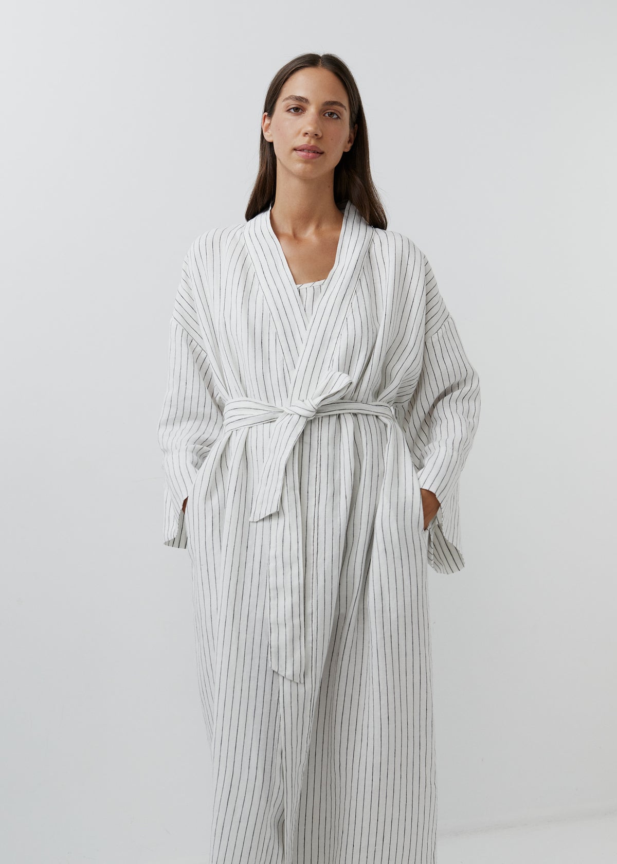 A woman wearing a long white linen robe with thin vertical black stripes, featuring long sleeves, a tie waist belt, and front pockets, set against a plain light gray studio background.