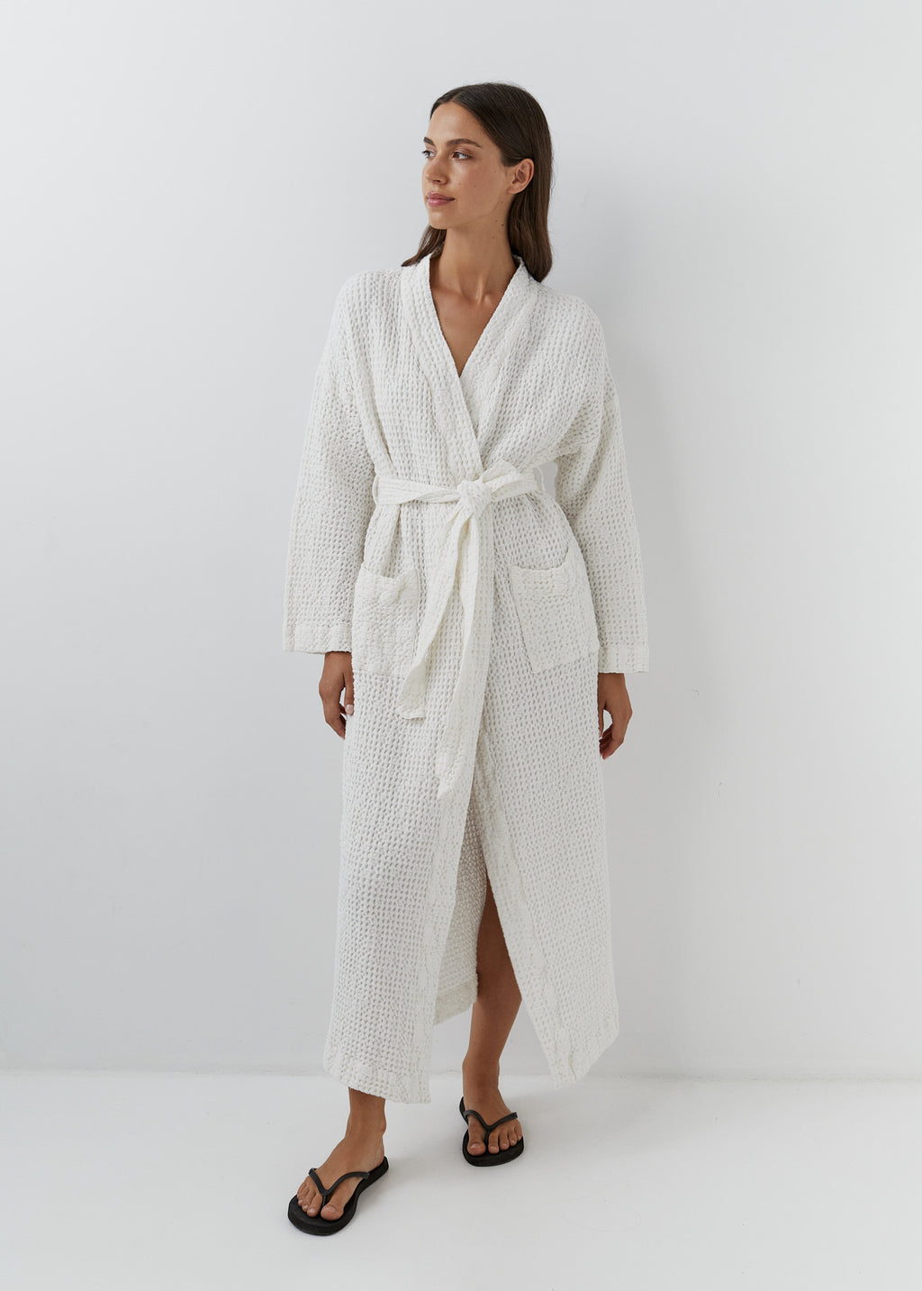 A woman modeling a long white waffle-weave bathrobe with a tie belt, standing against a plain white studio background.