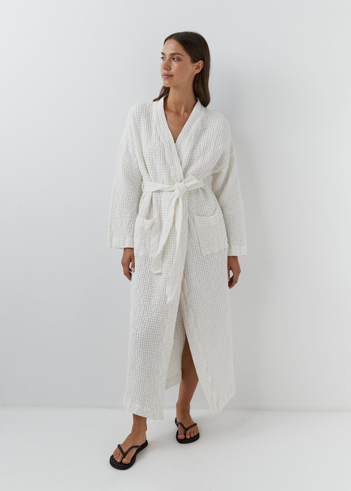 A woman modeling a long white waffle-weave bathrobe with a tie belt, standing against a plain white studio background.
