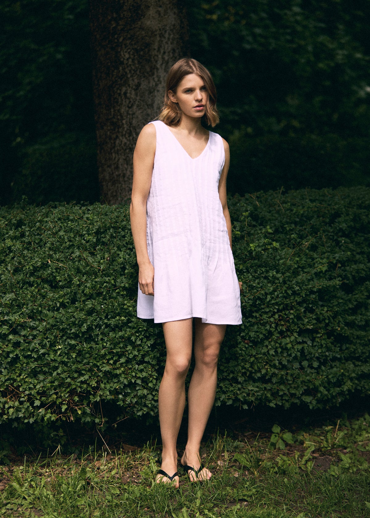 Woman wearing a white sleeveless linen mini dress with a V-neckline, relaxed A-line silhouette and above-the-knee length, standing outdoors in a green summer garden.
