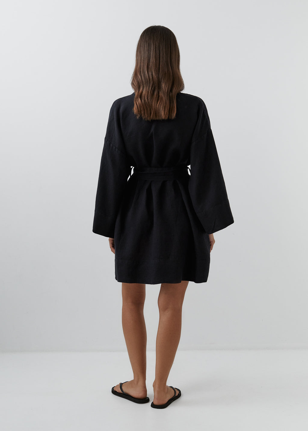 A woman modeling a short black linen kimono robe with a tie belt, shown from the back.
