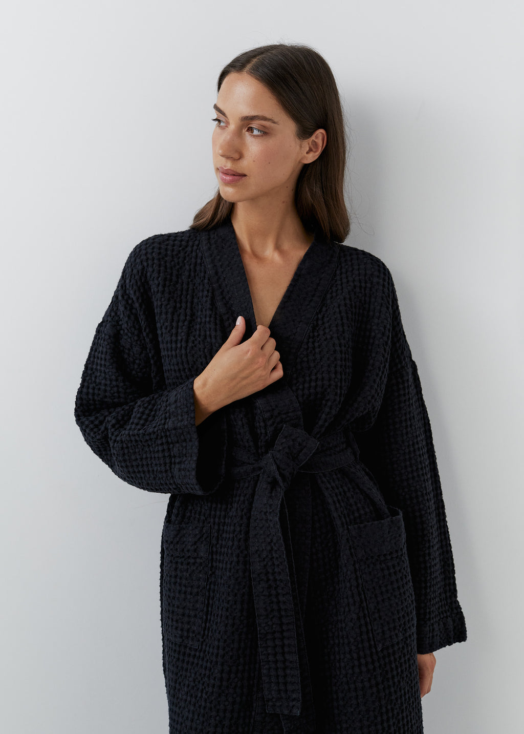 A woman wearing a black waffle linen cotton bathrobe, standing against a light gray wall.