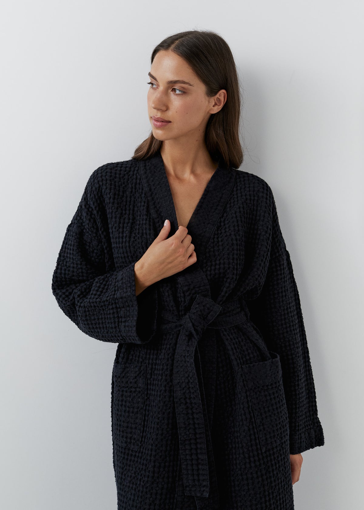 A woman wearing a black waffle linen cotton bathrobe, standing against a light gray wall.