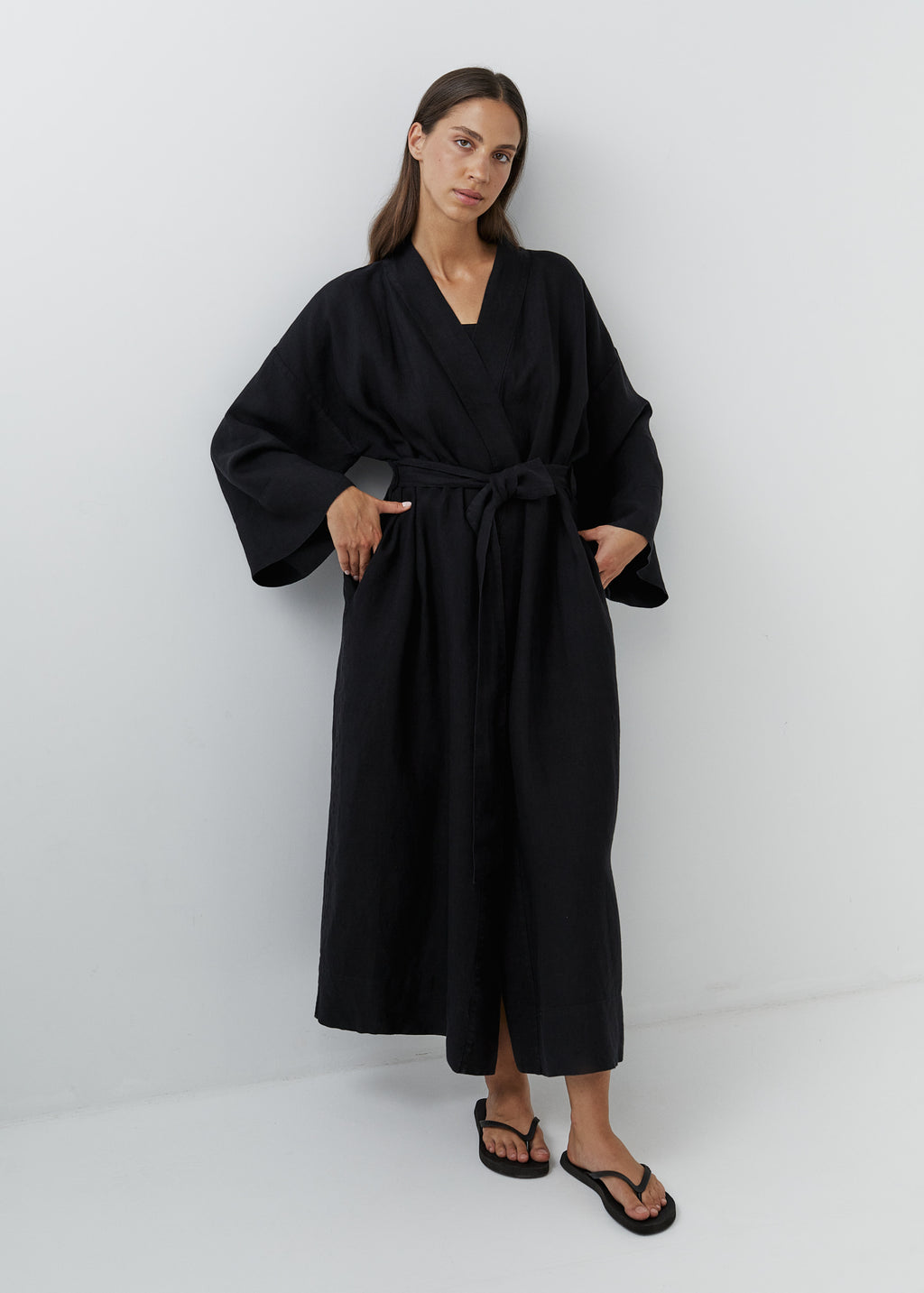 A woman modeling a long black linen kimono robe with wide long sleeves and a tie waist belt, standing against a plain light gray studio background.
