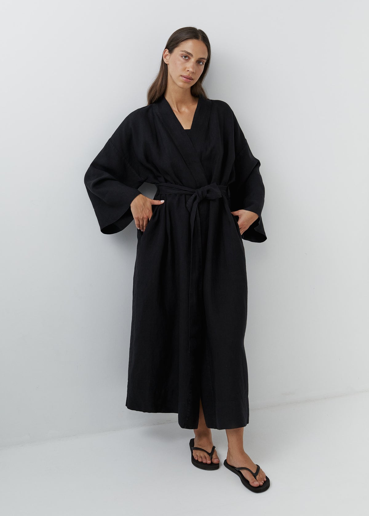 A woman modeling a long black linen kimono robe with wide long sleeves and a tie waist belt, standing against a plain light gray studio background.
