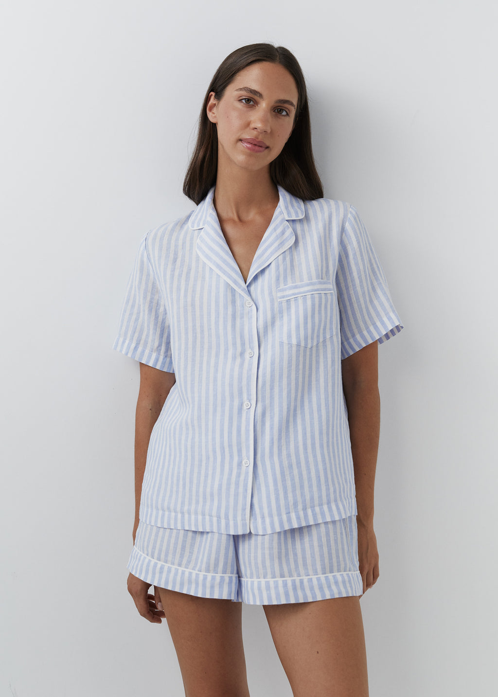 A woman modeling a light blue and white vertically striped short-sleeve pajama set with a button-front shirt and matching shorts against a plain light gray background.