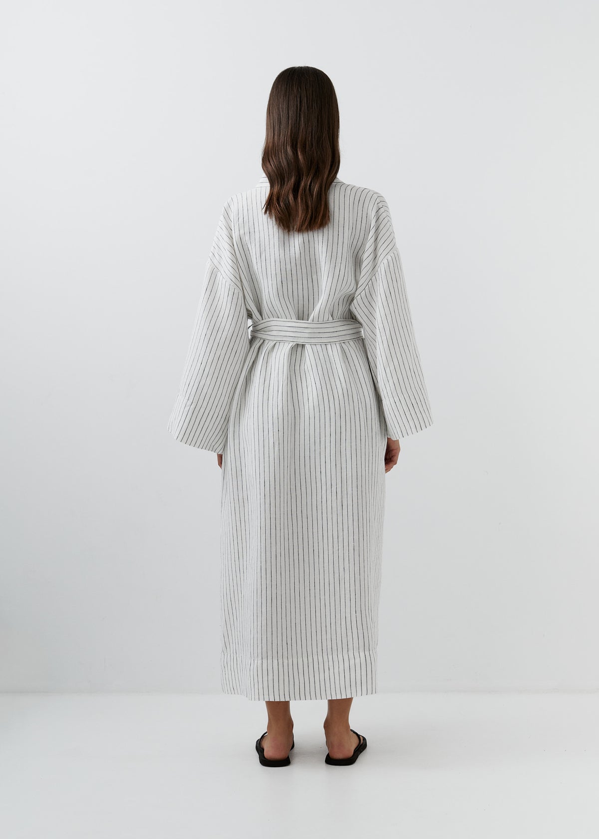 A woman viewed from behind wearing a long white robe with thin vertical black stripes and a matching waist tie, standing against a plain light gray studio background.