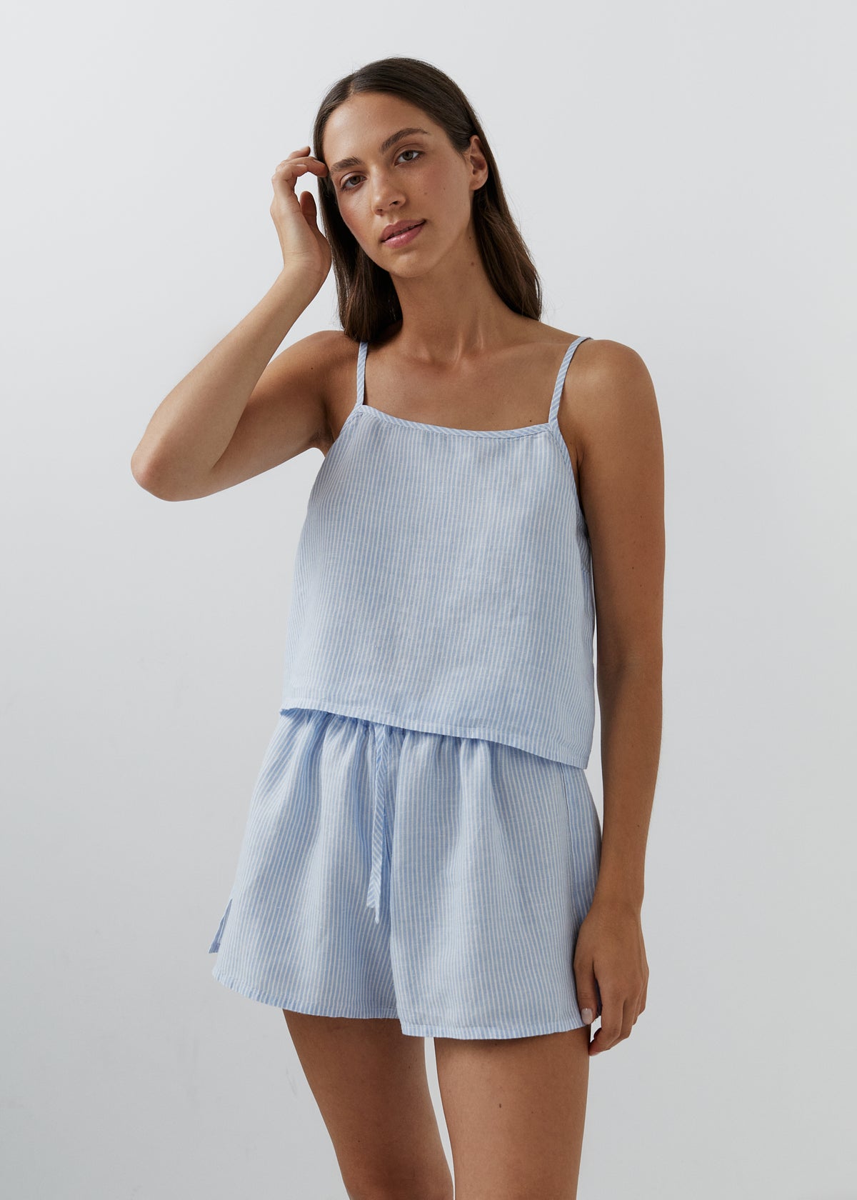 A woman modeling a light blue and white vertically striped two-piece set consisting of a spaghetti-strap camisole top and matching shorts, set against a plain light gray studio background.
