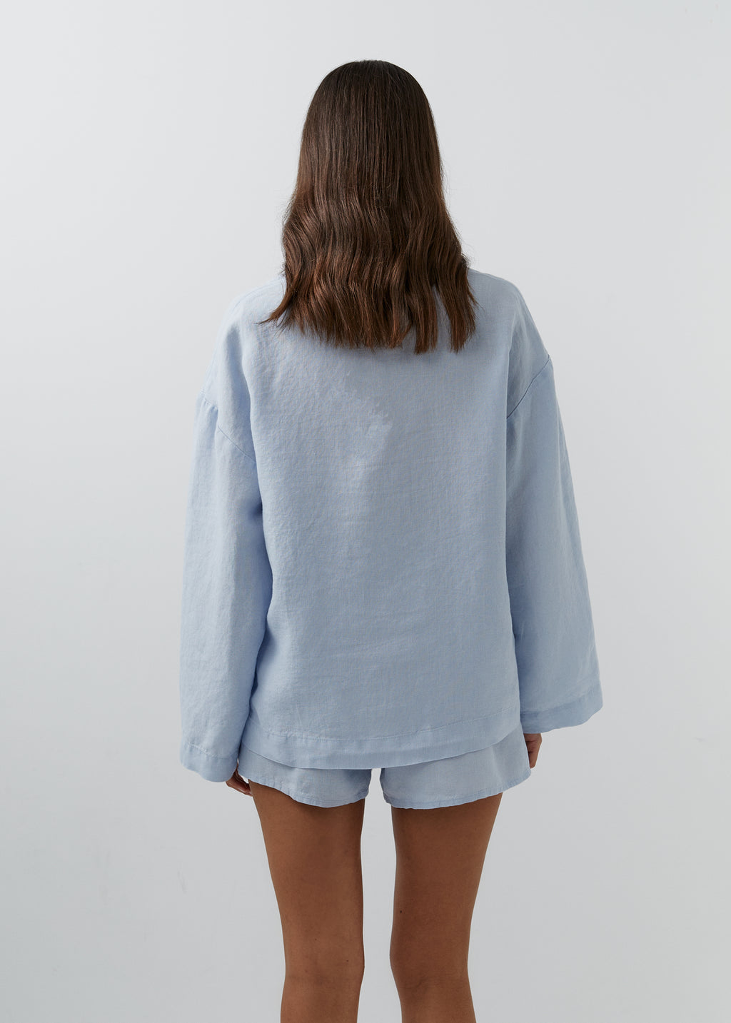 A woman viewed from behind wearing a light blue oversized long-sleeve top and matching light blue shorts against a plain light gray studio background.