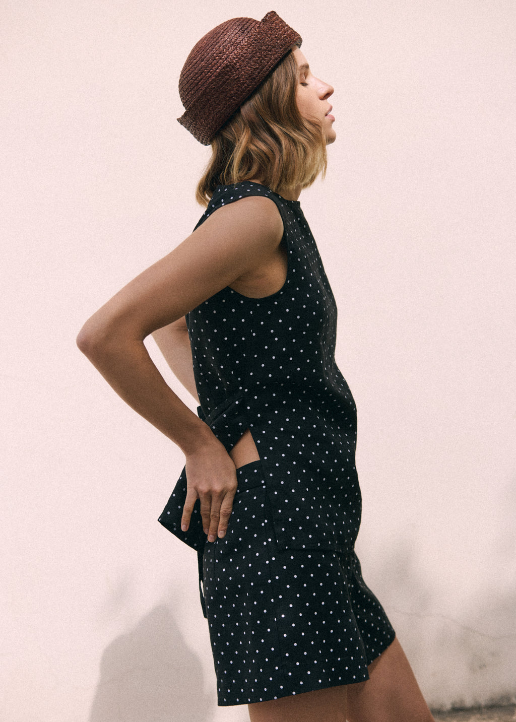 A woman wearing a sleeveless black top with white polka dots and matching shorts, paired with a brown woven hat, posing in profile against a light pink wall.