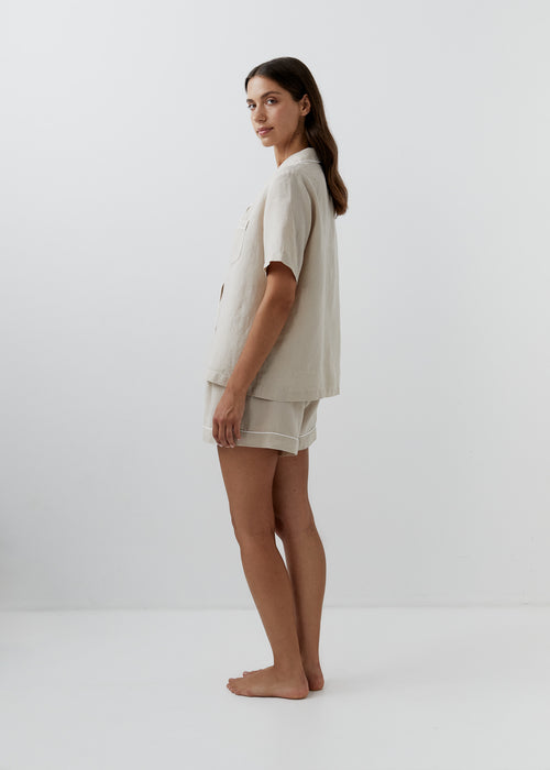 A woman stands in profile wearing a light beige short-sleeve button-front shirt and matching shorts set against a clean white studio background.