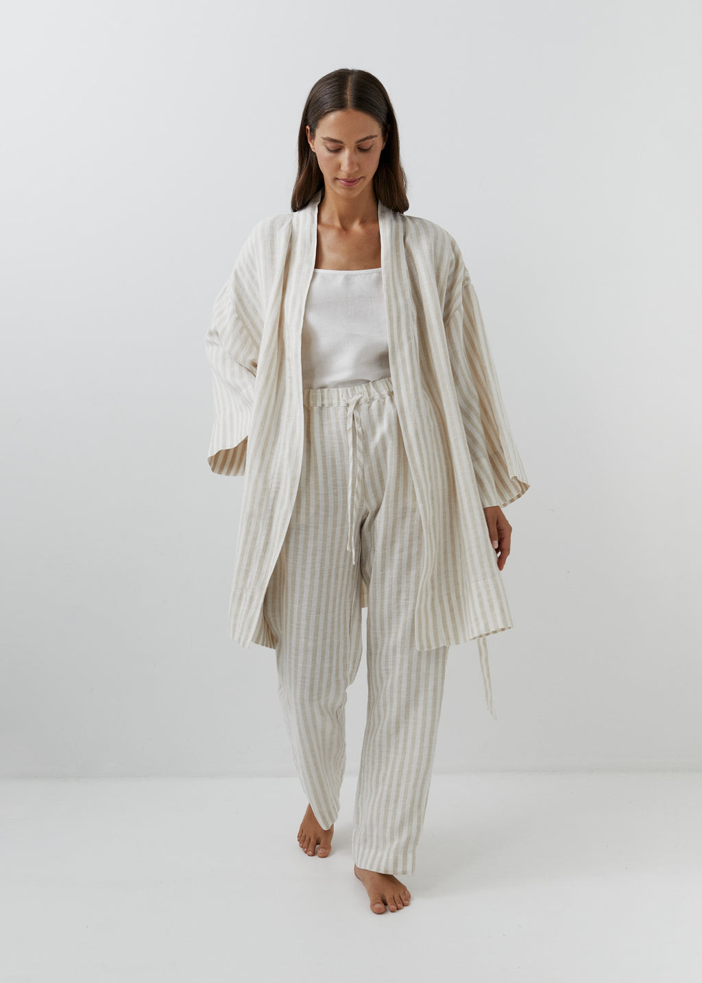 A woman modeling an open cream and white vertically striped linen robe with wide long sleeves, paired with matching striped linen trousers.