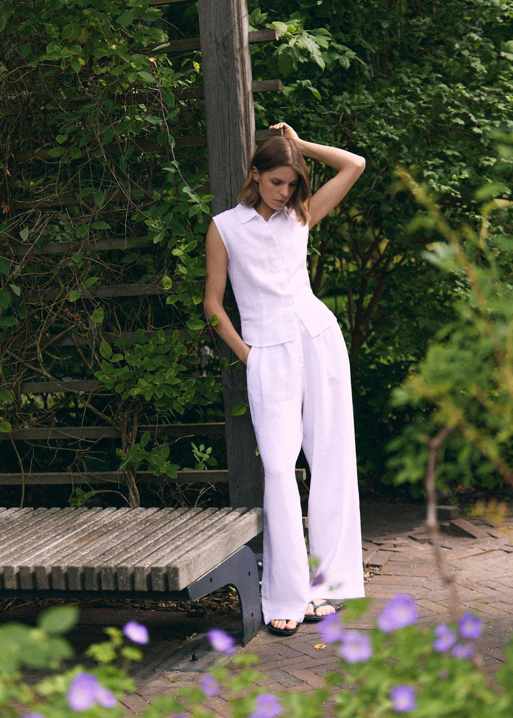 A woman models a white sleeveless collared top and matching wide-leg trousers in an outdoor garden setting with a wooden bench and green foliage.