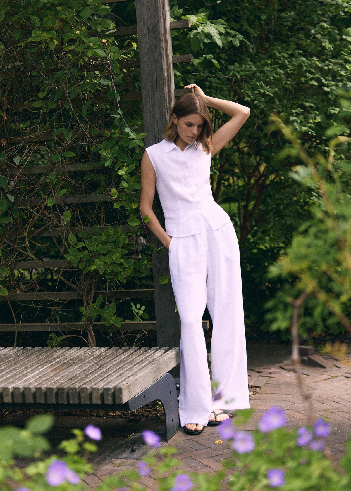 A woman models a white sleeveless collared top and matching wide-leg trousers in an outdoor garden setting with a wooden bench and green foliage.