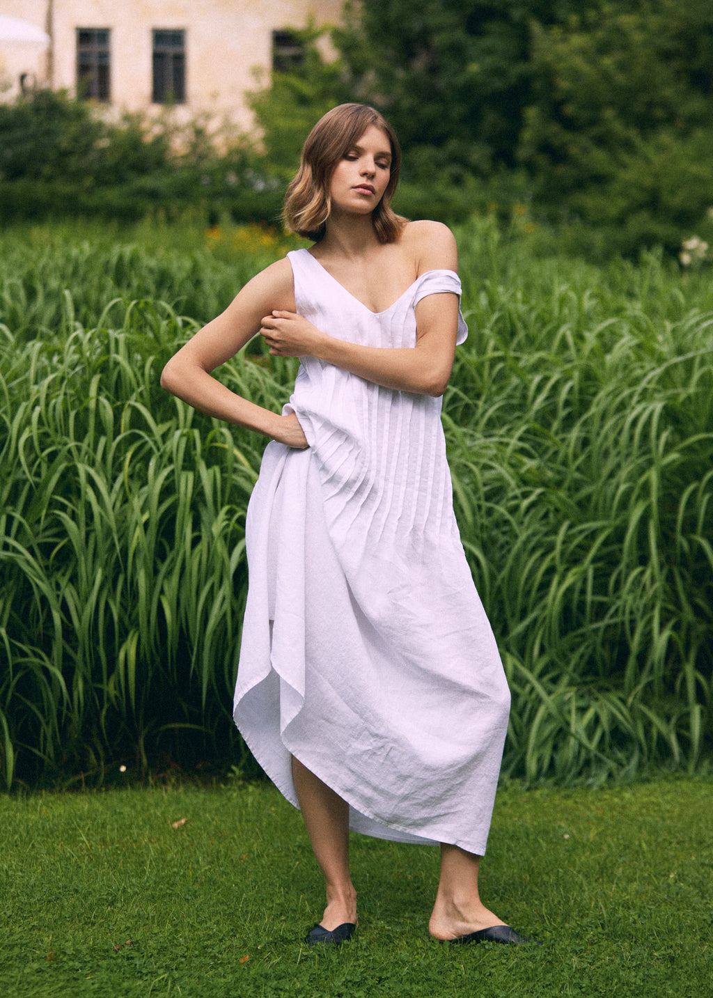 Woman posing in a garden wearing a white sleeveless linen maxi dress with a V-neckline, textured vertical pleats and a softly flowing skirt, paired with black flat shoes.